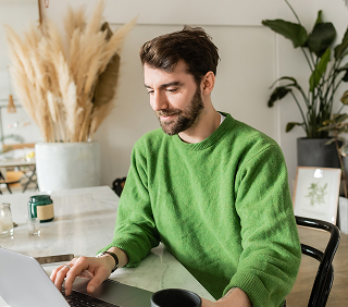 Man in green sweater works on laptop at a table, holding a coffee mug; cozy indoor setting with plants and dried pampas grass behind him.