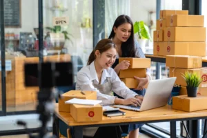 Two women in a small business pack orders at a desk with boxes; one manages spreadsheets on a laptop while the other holds packages.