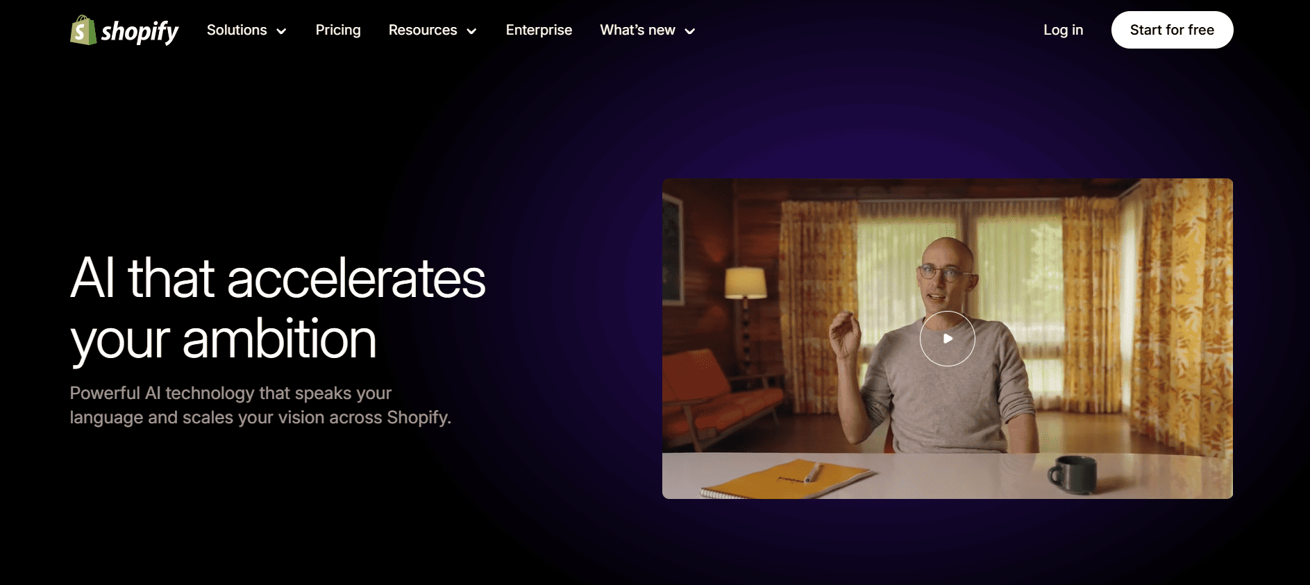 Shopify homepage with a banner reading "AI that accelerates your ambition." A person smiles, waving at their laptop in a cozy room.