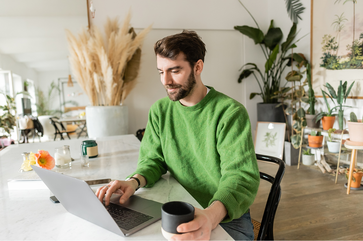 A man in a green sweater uses a laptop at a sunlit table, holding a black mug in a cozy, modern room filled with plants.